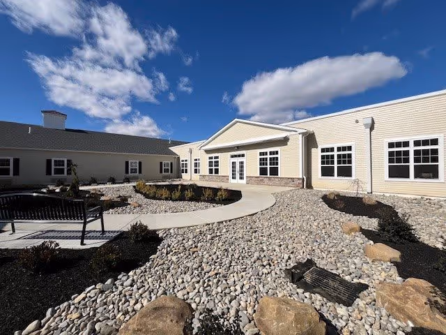 Sunny courtyard with pebble landscaping, a curved concrete walkway and benches in front of a low beige senior living building under a blue sky.