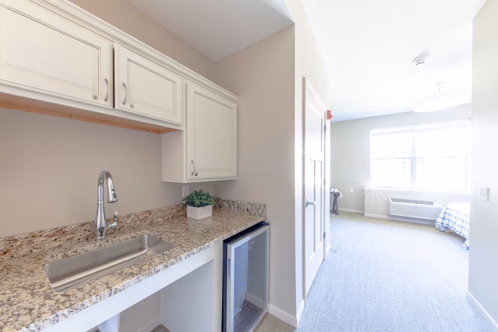 Interior view of a small kitchenette area with a granite countertop, stainless steel sink, white cabinets, and a mini refrigerator. The kitchenette opens into a bright room with a large window, carpeted floor, and a bed partially visible on the right side.