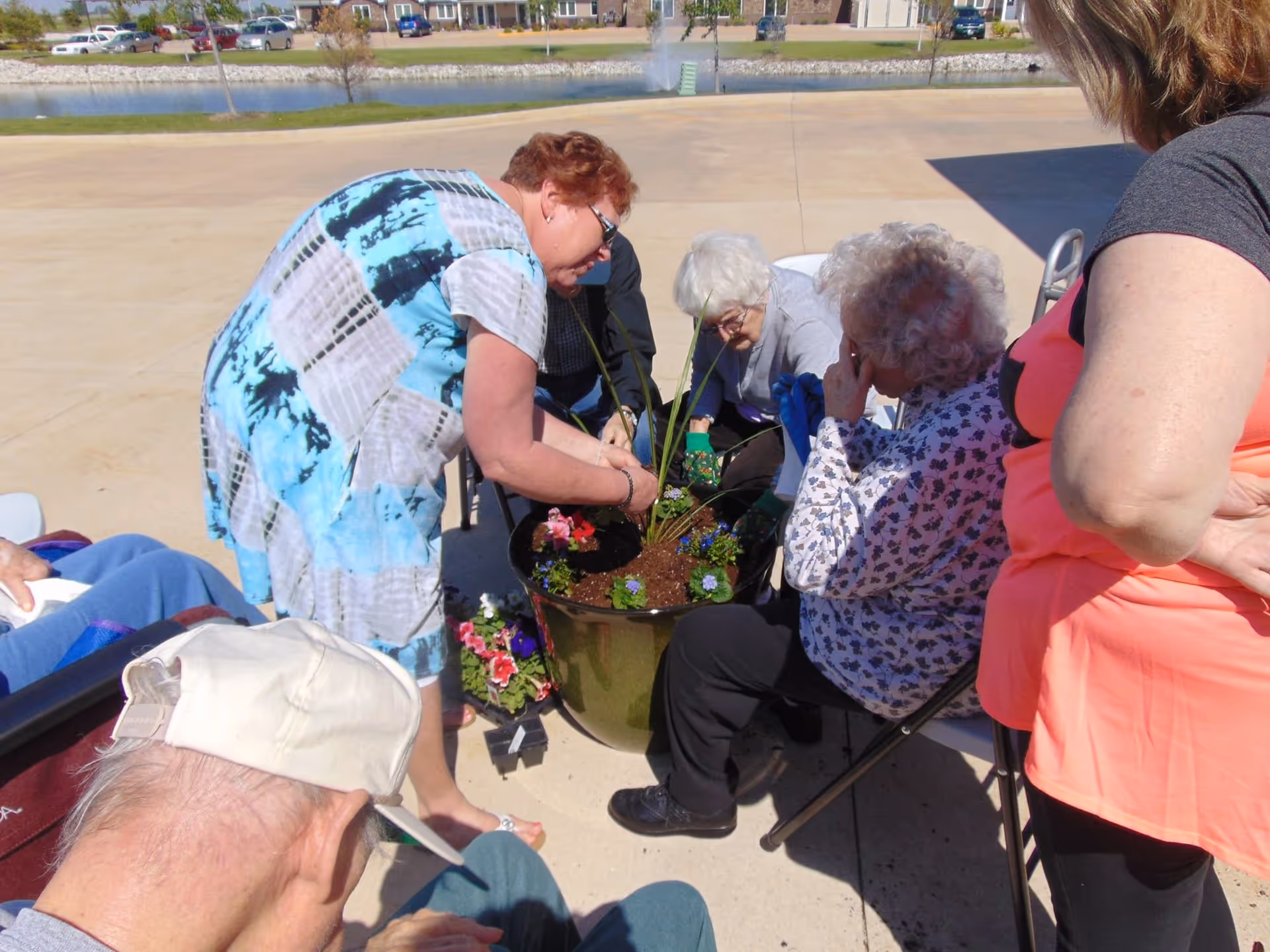 A group of elderly people and a caregiver are gathered outdoors around a large planter pot, engaging in a gardening activity. The caregiver is arranging plants in the pot while the elderly participants watch and assist. The setting is a paved area near a pond with a fountain and residential buildings in the background.