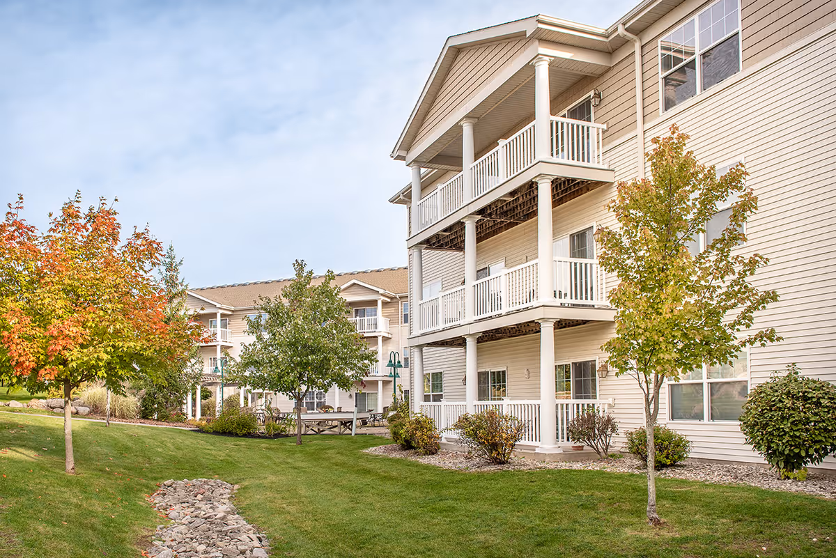 Exterior view of a multi-story residential building with beige siding and white balconies. The building is surrounded by a well-maintained lawn with small trees, some showing autumn colors, and landscaping with rocks and bushes under a partly cloudy sky.