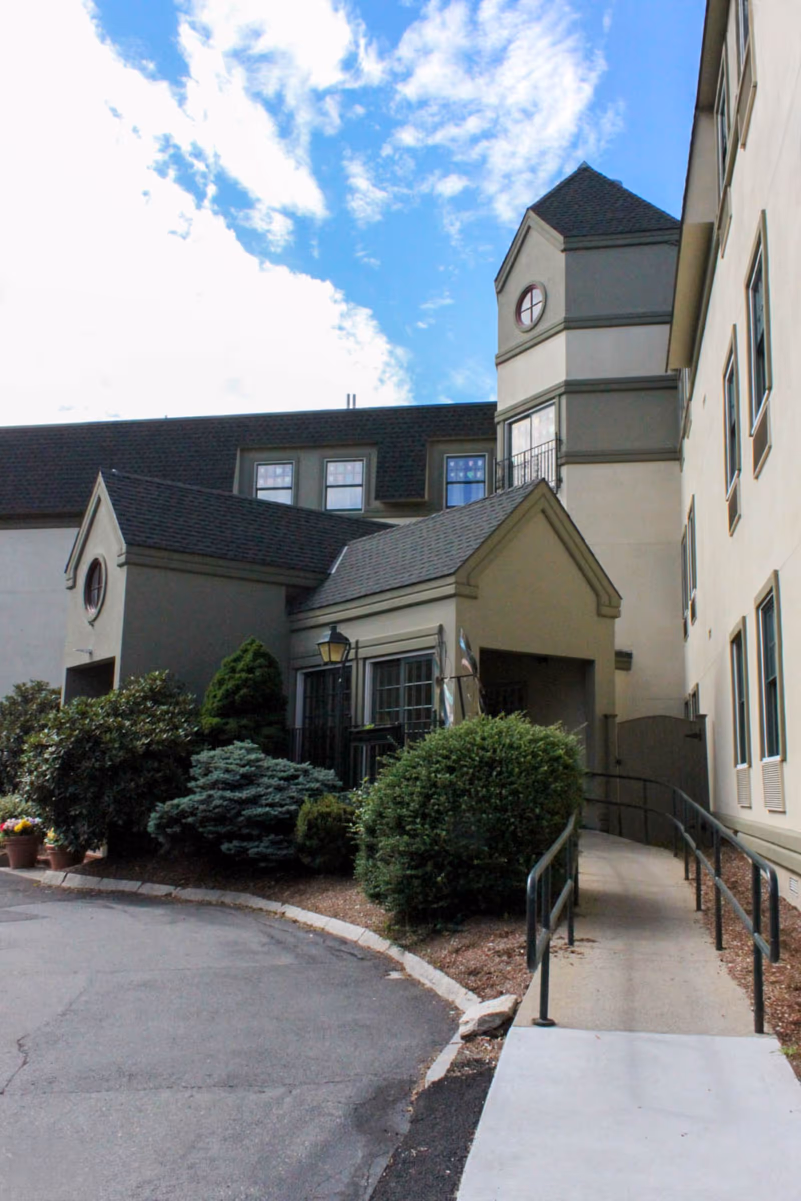 Exterior view of Brandon Woods of New Bedford facility showing a beige building with multiple windows, a small tower with a circular window, a sloped roof, and a ramp with handrails leading to an entrance. There are bushes and plants along the building and a partly cloudy blue sky above.