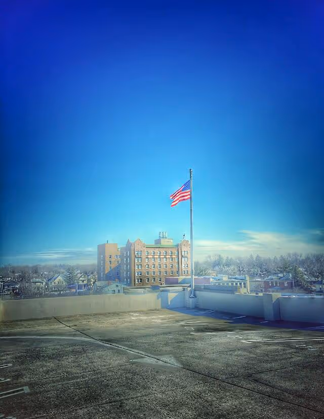 View of a rooftop parking area with an American flag on a flagpole in the center. In the background, there is a multi-story brick building and a neighborhood with trees under a clear blue sky.
