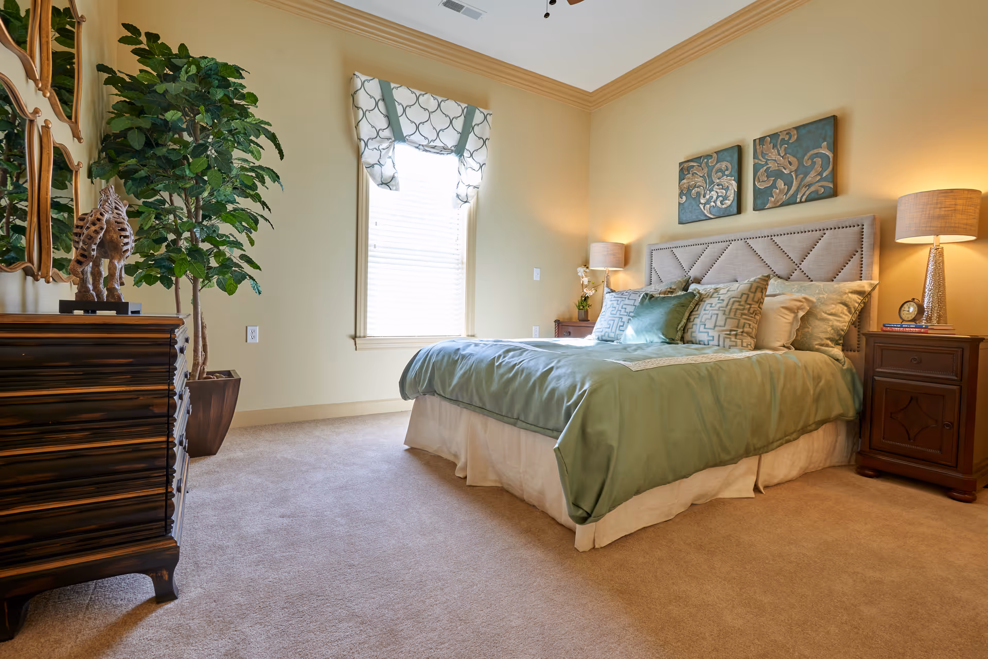 Bright bedroom featuring a large bed with green bedding, matching nightstands and lamps, a dresser, wall art, and a potted plant.