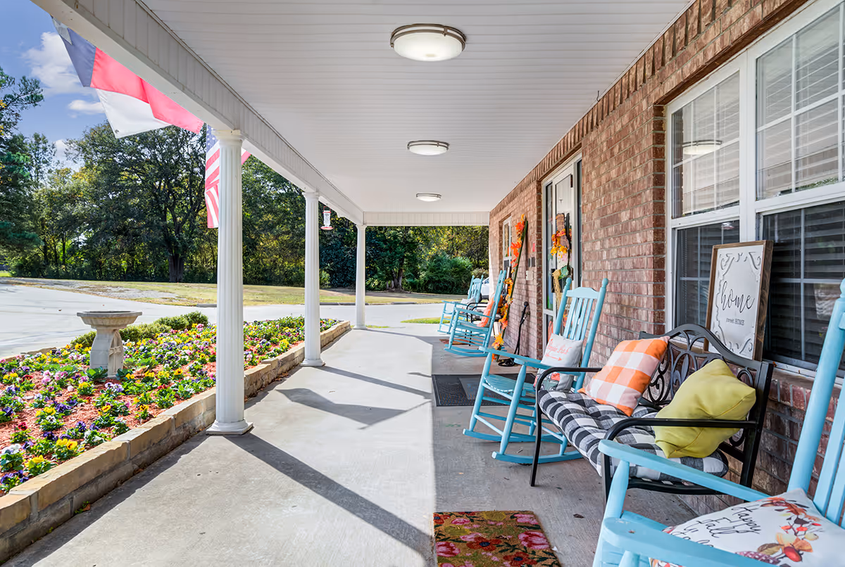 A covered porch area with light blue rocking chairs and a black metal bench with colorful cushions. The porch has white columns and a flower bed with vibrant flowers along the edge. There are two flags hanging from the porch ceiling, and the building has a brick exterior with windows and a door decorated with fall-themed wreaths.