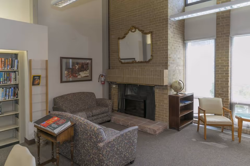 A seating area with patterned sofas and chairs facing a brick fireplace with a mirror, plus a bookshelf and tall windows.