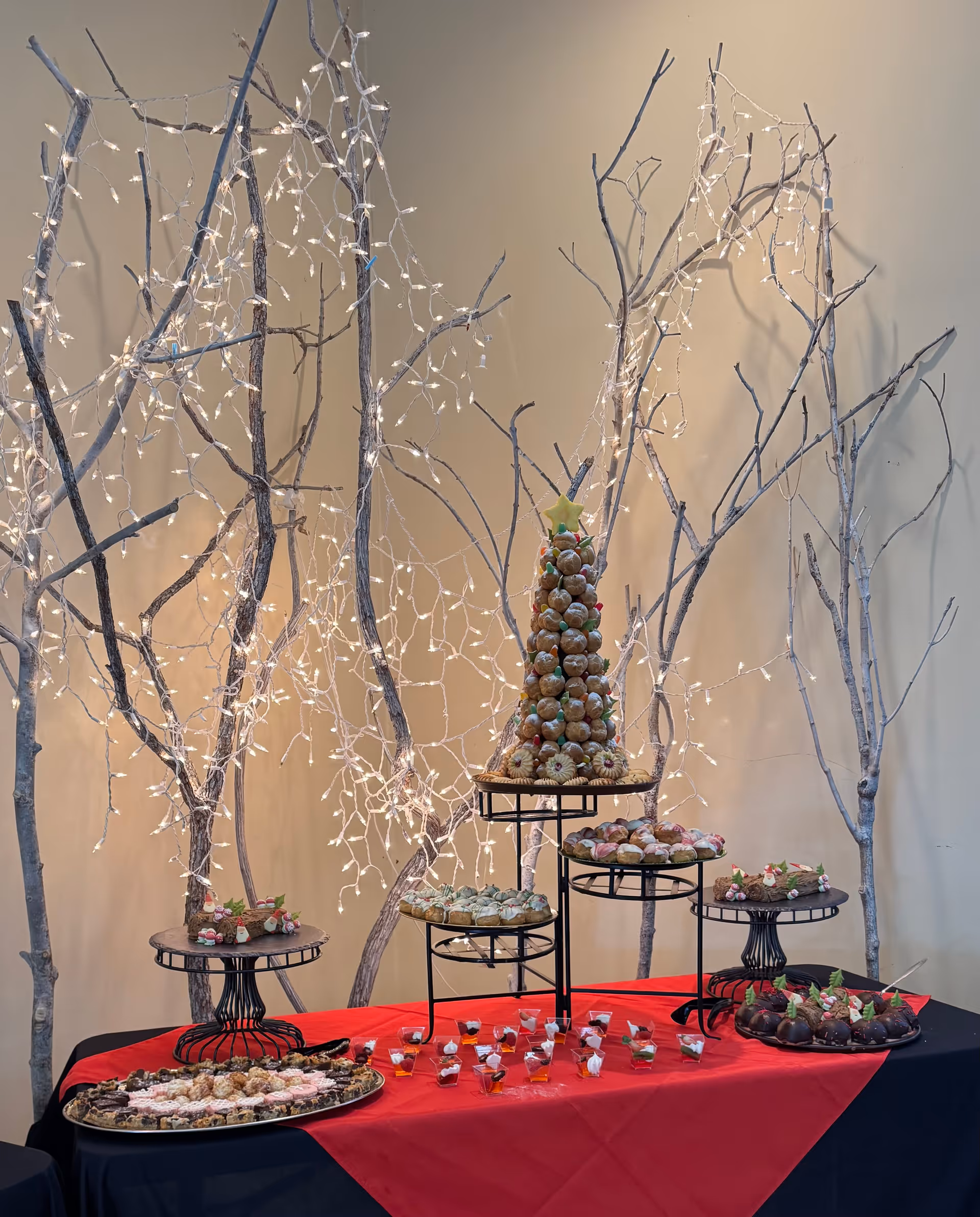 A festive dessert display featuring a croquembouche tower decorated with a star on top, surrounded by various pastries and small dessert cups on a table covered with a red and black tablecloth. Behind the table are bare tree branches adorned with white string lights against a beige wall.