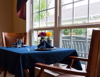 A dining table covered with a dark blue tablecloth set with folded napkins, glasses, and silverware, positioned near a large window with a view of an outdoor patio and greenery. Two wooden chairs are placed at the table.
