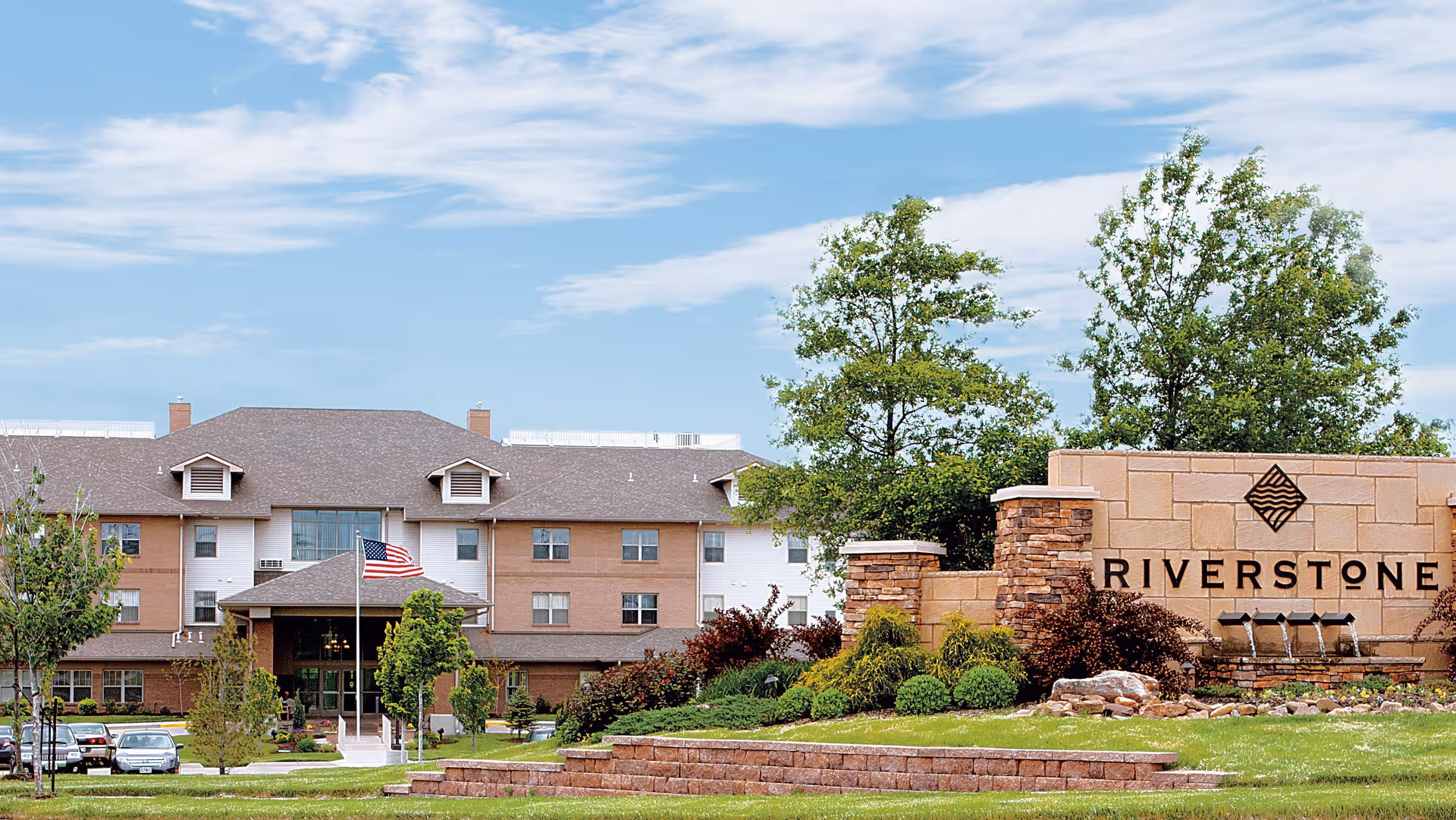 Front exterior of the Riverstone Retirement Community showing the main building, landscaped entrance sign, fountain, and an American flag.