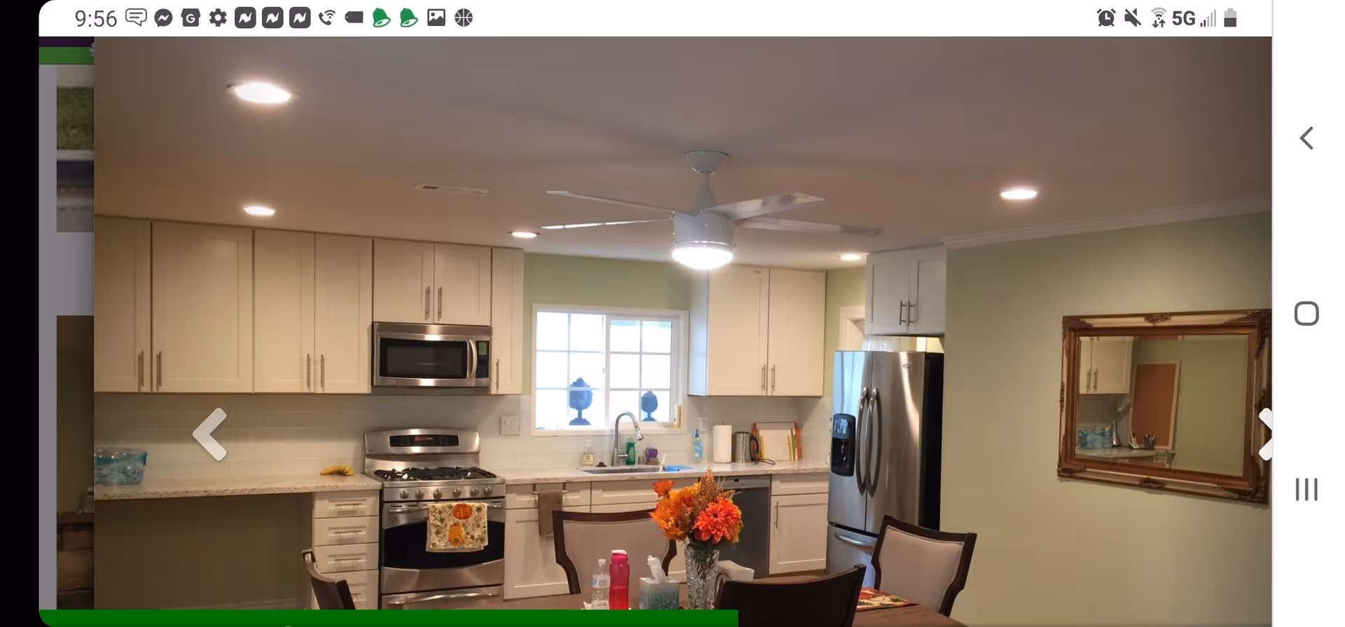 A modern kitchen with white cabinets, stainless steel appliances including a stove, microwave, and refrigerator. There is a window above the sink letting in natural light. A ceiling fan with a light is mounted on the ceiling. In the foreground, there is a dining table with chairs and a vase of colorful flowers on it. A large mirror hangs on the wall to the right.