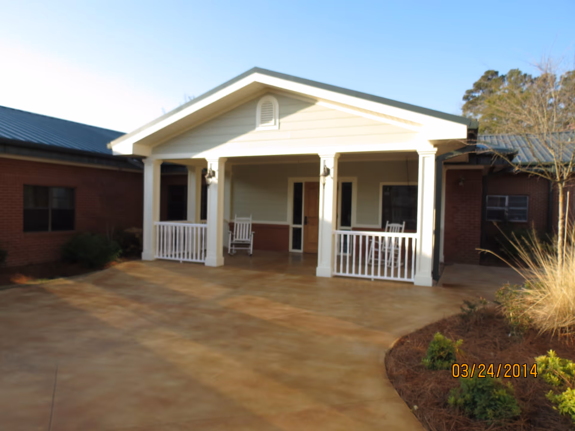 Front exterior view of a single-story building with a covered porch featuring white railings and rocking chairs, surrounded by a paved area and some landscaping with bushes and trees under a clear sky.