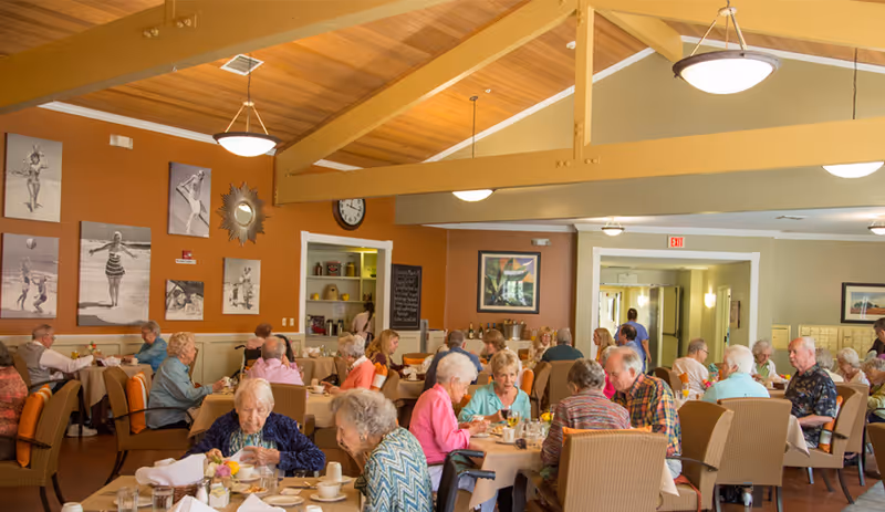 A dining room in an assisted living community with elderly residents seated at tables enjoying meals. The room has warm orange and beige walls, wooden ceiling beams, and black and white photos on one wall. Several round tables are set with white tablecloths, dishes, and flowers. The atmosphere is bright and social.
