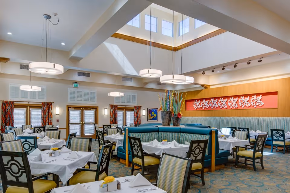 Bright and spacious dining room with multiple tables covered in white tablecloths, set with napkins and silverware. The room features teal cushioned booth seating, patterned chairs, large windows with orange patterned curtains, modern hanging light fixtures, and a skylight allowing natural light to fill the space. Decorative plants and artwork adorn the walls.