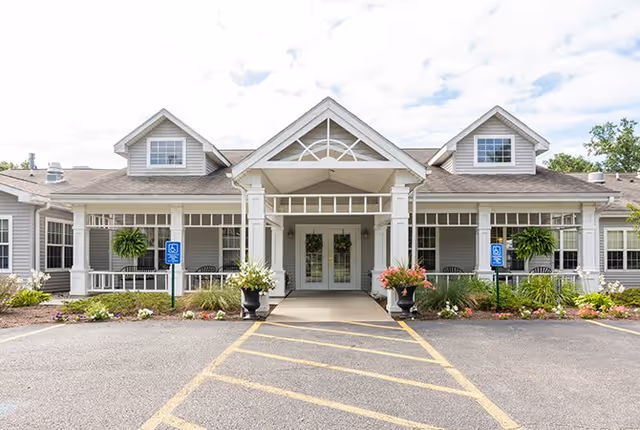 Front exterior view of a single-story building with a covered entrance supported by white columns. The building has gray siding, multiple windows, and two dormer windows on the roof. There are handicap parking signs and landscaped flower beds on either side of the entrance.