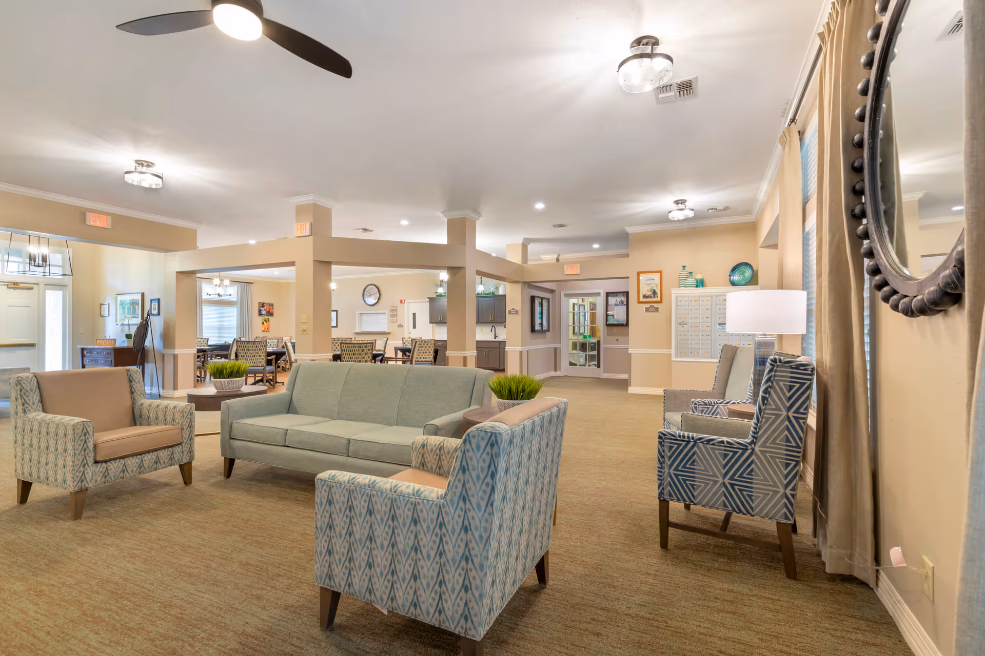 A spacious and well-lit common area in a senior living facility featuring a seating arrangement with a green sofa, two patterned armchairs, and a beige armchair. The room has beige walls, carpeted floors, ceiling lights, a ceiling fan, and decorative elements including a round mirror and table lamps. In the background, there are dining tables and chairs, a bulletin board, and a doorway leading to other parts of the facility.