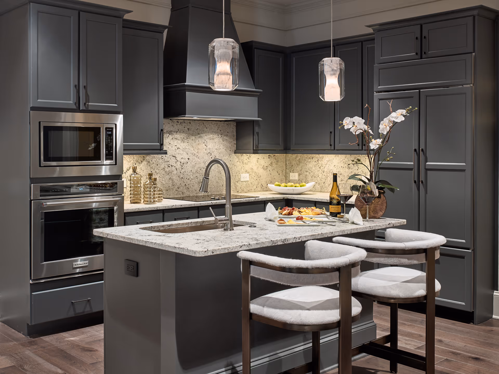 Modern kitchen with dark gray cabinets, a granite countertop island with a built-in sink, two cushioned bar stools, stainless steel microwave and oven, pendant lights, and a decorative orchid plant. A bottle of wine and glasses are placed on the island along with a plate of appetizers.