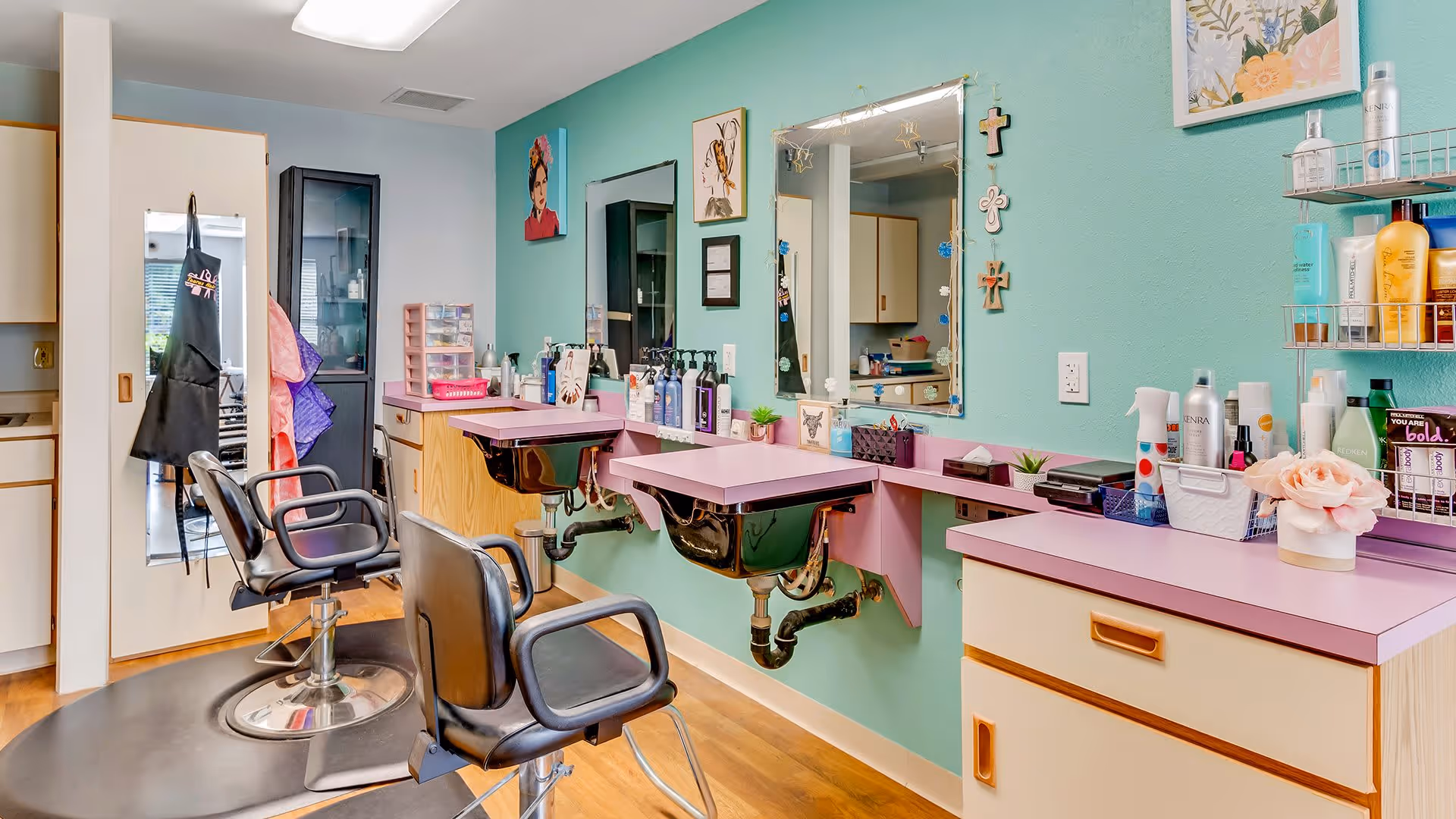 Interior view of a salon area in a senior living facility with two black salon chairs in front of pink countertops and sinks mounted on a teal wall. The wall has mirrors, hair care products, and decorative items including small framed pictures and crosses. There is a cabinet and a door with a hanging black apron on the left side.