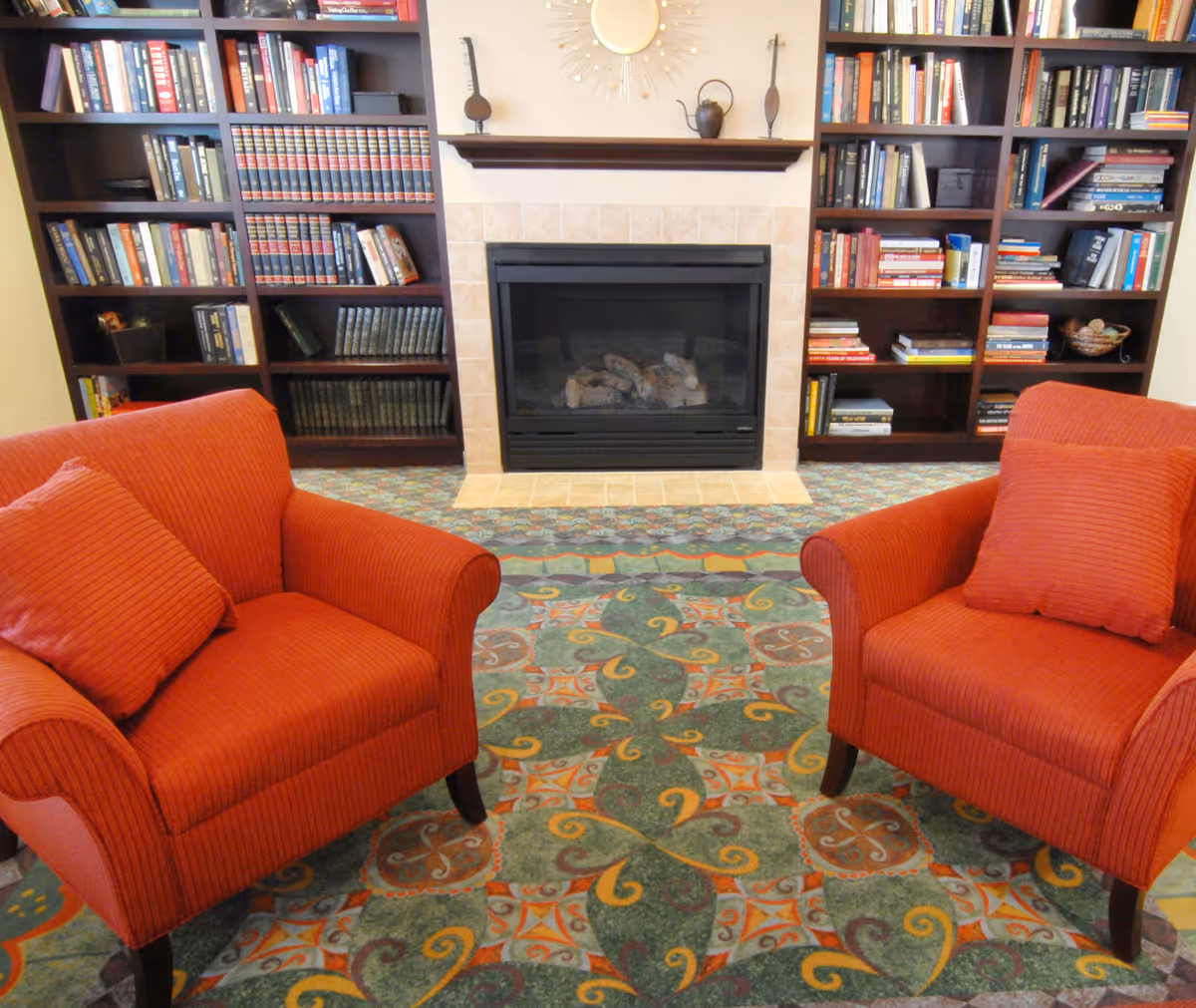 Two red armchairs facing a fireplace flanked by bookshelves in a carpeted sitting area.