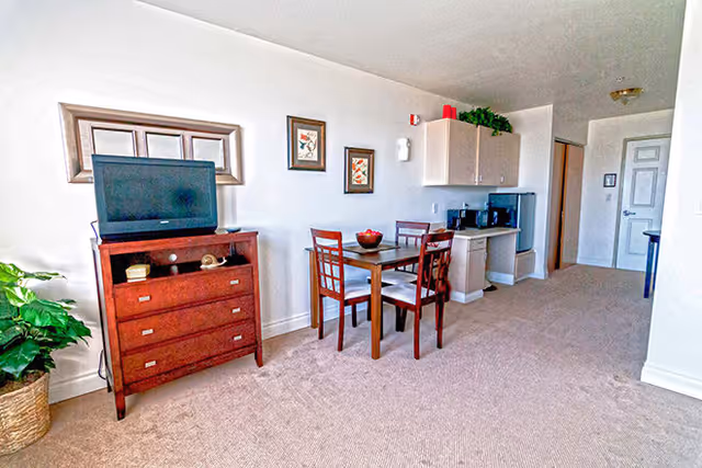 Interior view of a senior living facility room featuring a small dining table with four chairs, a wooden dresser with a television on top, framed artwork on the wall, a small kitchenette area with cabinets, a microwave, and a mini refrigerator, and a carpeted floor.