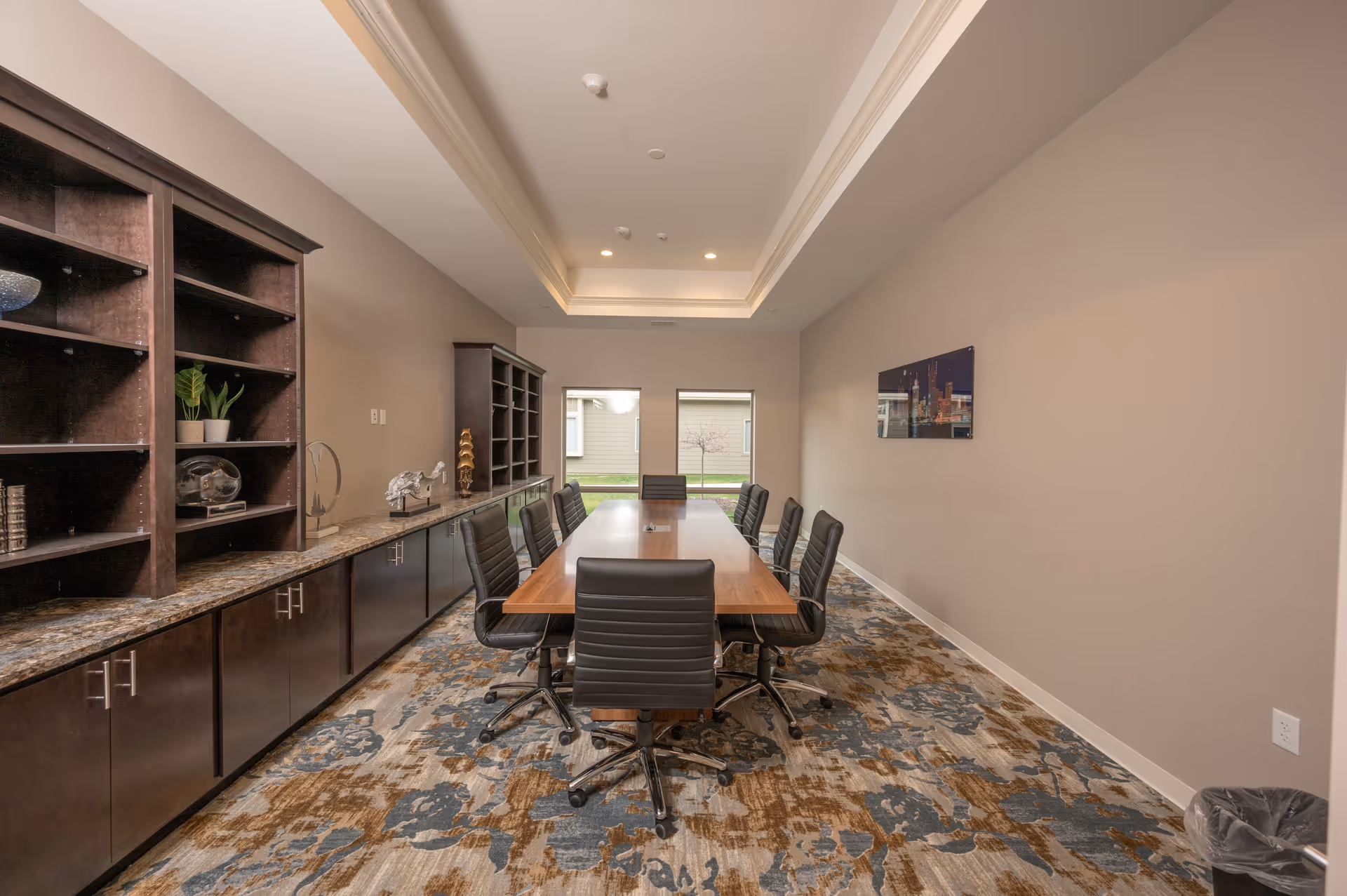 A modern conference room with a long wooden table surrounded by black office chairs on wheels. The room has a patterned carpet in shades of blue and brown, beige walls, and a recessed ceiling with built-in lighting. On the left side, there are dark wooden cabinets and shelves with decorative items and plants. Two windows at the far end provide natural light and a view of the outside. A framed picture hangs on the right wall, and a trash can is visible in the bottom right corner.