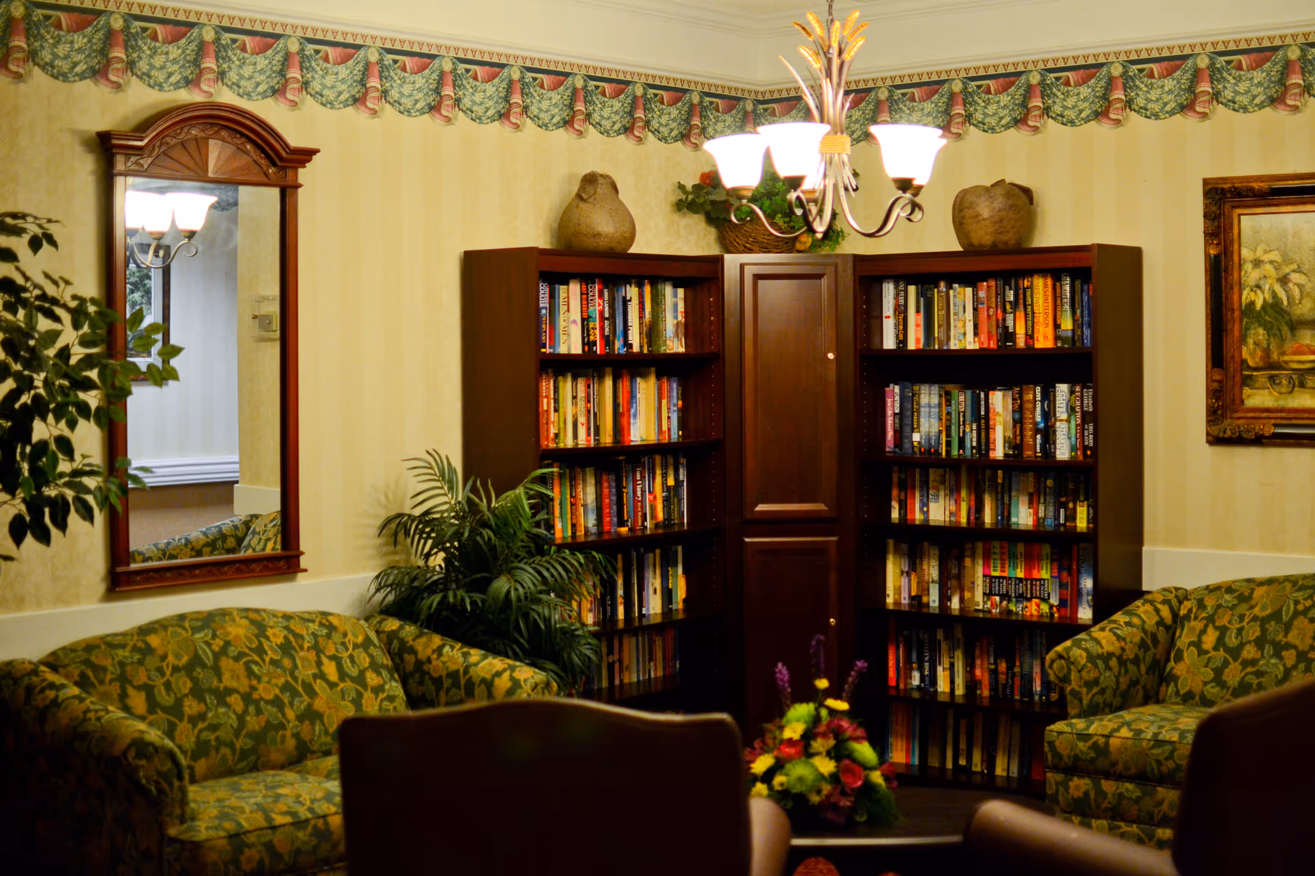 A cozy living room area with two green floral patterned sofas facing each other, a dark wooden bookshelf filled with books in the corner, a decorative mirror on the wall, a chandelier with five lights hanging from the ceiling, and a floral arrangement on a table in the foreground. The walls have a yellow striped wallpaper with a green and red patterned border near the ceiling.