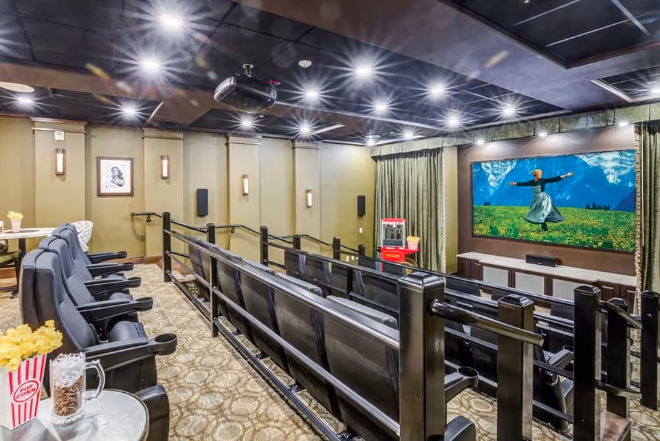 Interior view of a small movie theater room with multiple rows of black reclining seats, a large screen showing a scene from The Sound of Music, a popcorn machine, and tables with popcorn and snacks. The room has carpeted floors, wall sconces, and a dark ceiling with recessed lighting.