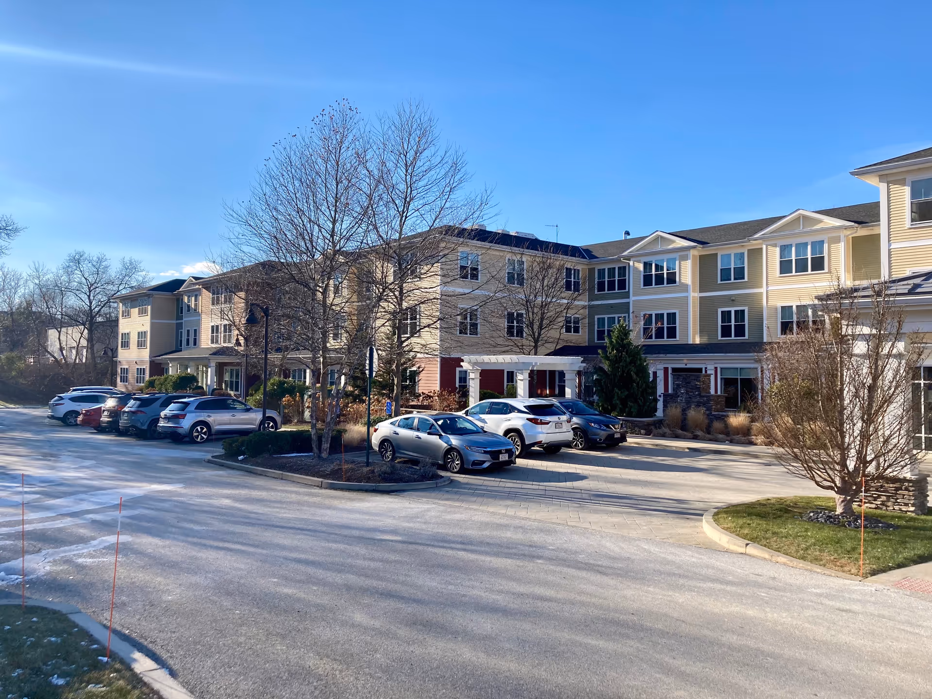 Front exterior of a multi-story senior living building with parked cars, bare trees, and a clear blue sky.