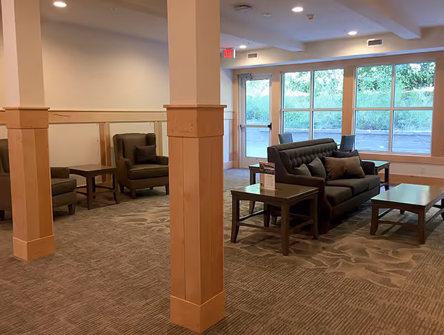Interior view of a common area in a senior living facility with carpeted floors, wooden pillars, a dark cushioned sofa with pillows, two armchairs, multiple wooden side tables, large windows letting in natural light, and a glass door leading outside.