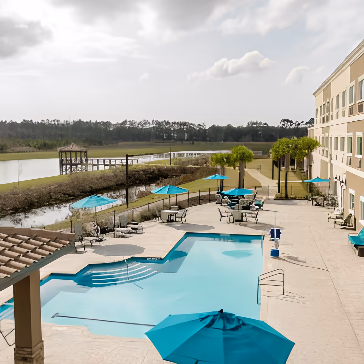 Outdoor swimming pool area at Discovery Village At Deerwood Assisted Living & Memory Care with turquoise umbrellas, lounge chairs, and a view of a lake and trees in the background.