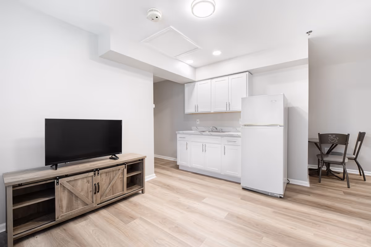 Small open living area with a TV on a wooden console, a kitchenette with white cabinets and a refrigerator, and a small dining table.