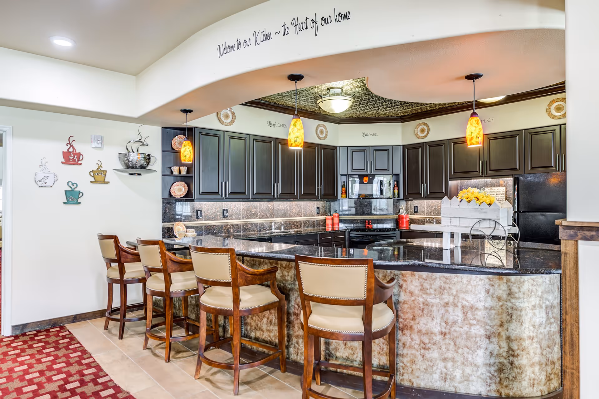 A modern kitchen area with a curved granite countertop bar and four cushioned wooden bar stools. The kitchen features dark wood cabinets, a black refrigerator, and a black stove. Three pendant lights with orange-yellow shades hang above the bar. The wall above the kitchen arch has the phrase 'Welcome to our Kitchen ~ the Heart of our home' written on it. Decorative coffee cup wall art is displayed on the left wall, and a red patterned carpet runs along the floor in front of the bar.