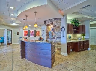 Interior view of a senior living facility reception area with a curved wooden front desk, pendant lights hanging from the ceiling, framed artwork on the wall, a stone accent wall, and a kitchenette area with cabinets and under-cabinet lighting.