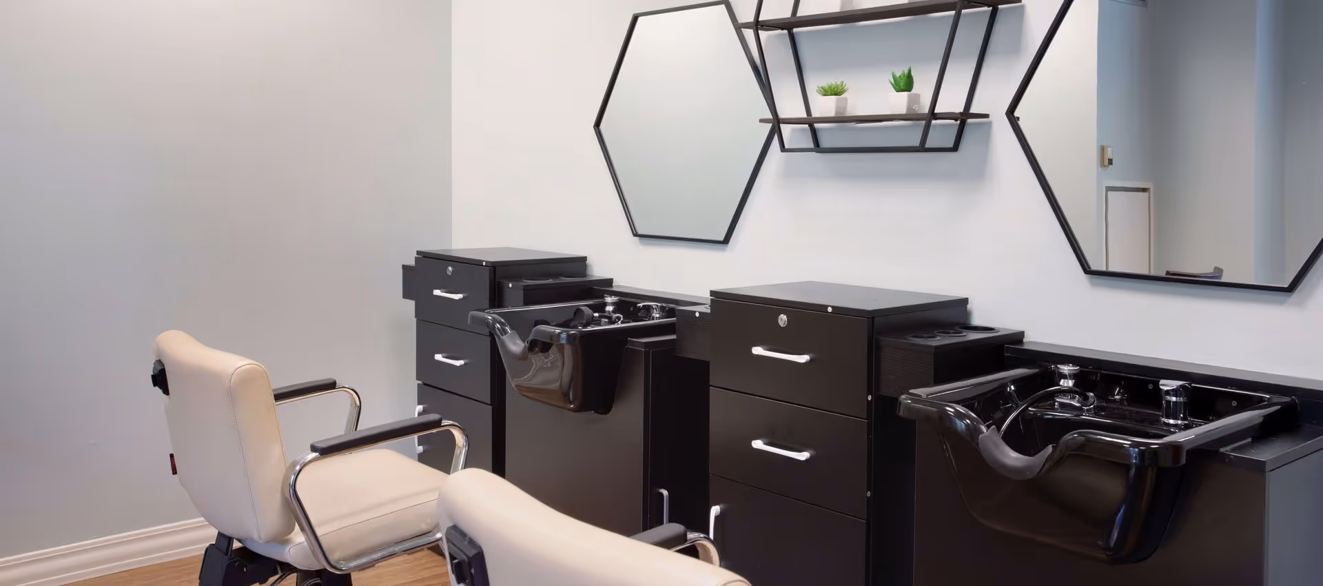 Interior view of a salon area with two beige salon chairs facing black hair washing stations with sinks and mirrors on the wall. A small shelf with two small potted plants is mounted above one of the sinks.