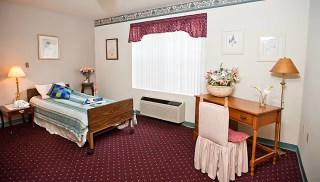 A cozy assisted living bedroom with a single bed covered in a blue and green striped blanket, a wooden desk with a floral arrangement, a chair with a pink skirted cover, a floor lamp, and framed artwork on the walls. The room has a maroon carpet with a small pattern and a window with vertical blinds and a maroon valance.