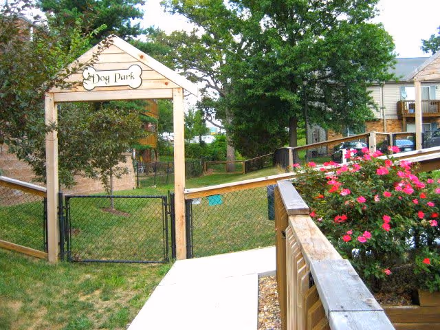 A fenced dog park area with a wooden archway labeled 'Dog Park' at the entrance. The park is surrounded by grass and trees, with residential buildings visible in the background. A wooden railing with blooming pink flowers is on the right side of the image.