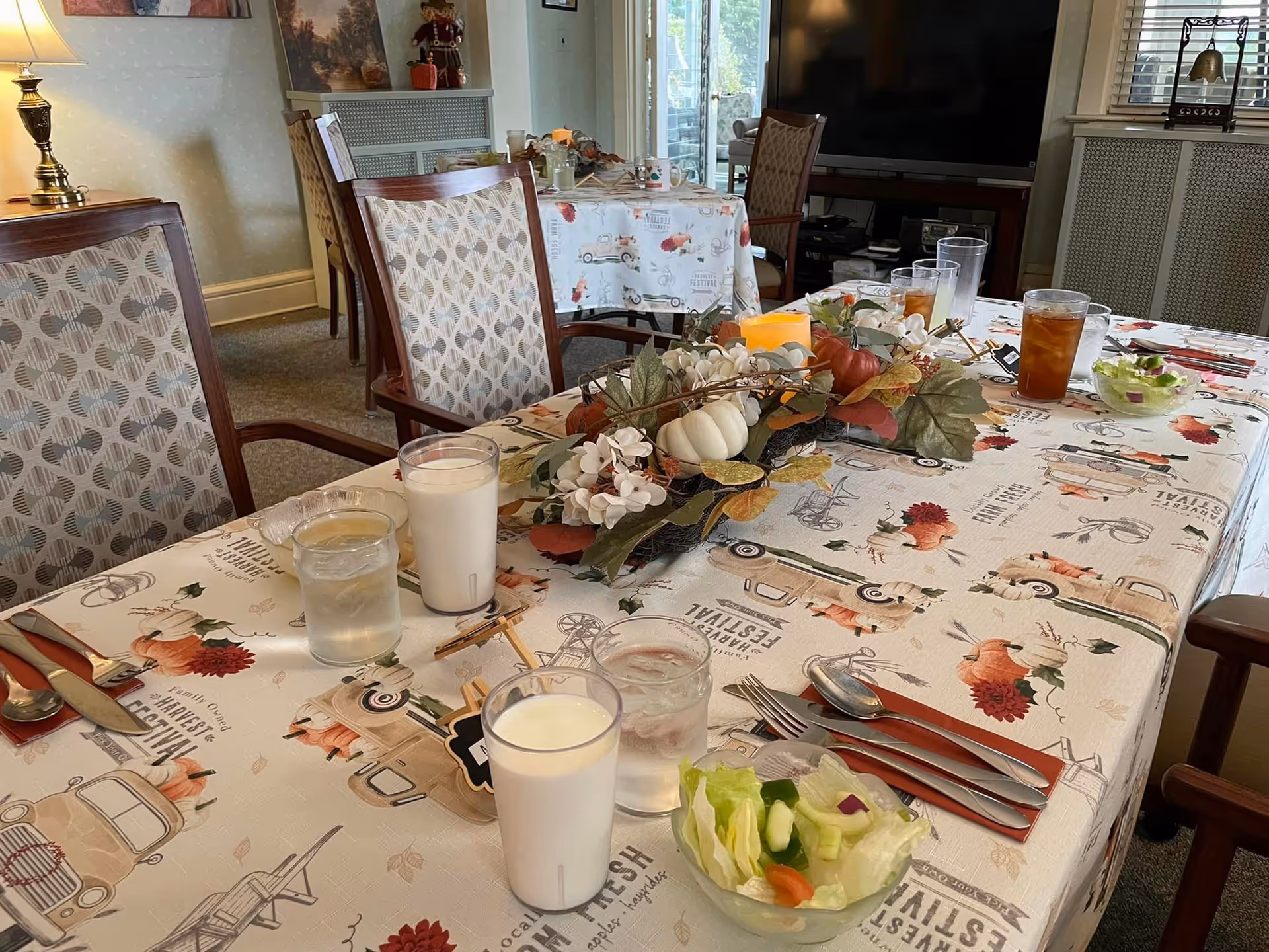 A dining table set with glasses of milk, water, iced tea, and small bowls of salad. The table is covered with a festive tablecloth featuring autumn and harvest-themed designs, and a centerpiece with artificial pumpkins, flowers, and leaves. The room has patterned chairs, a lamp, and a television in the background.