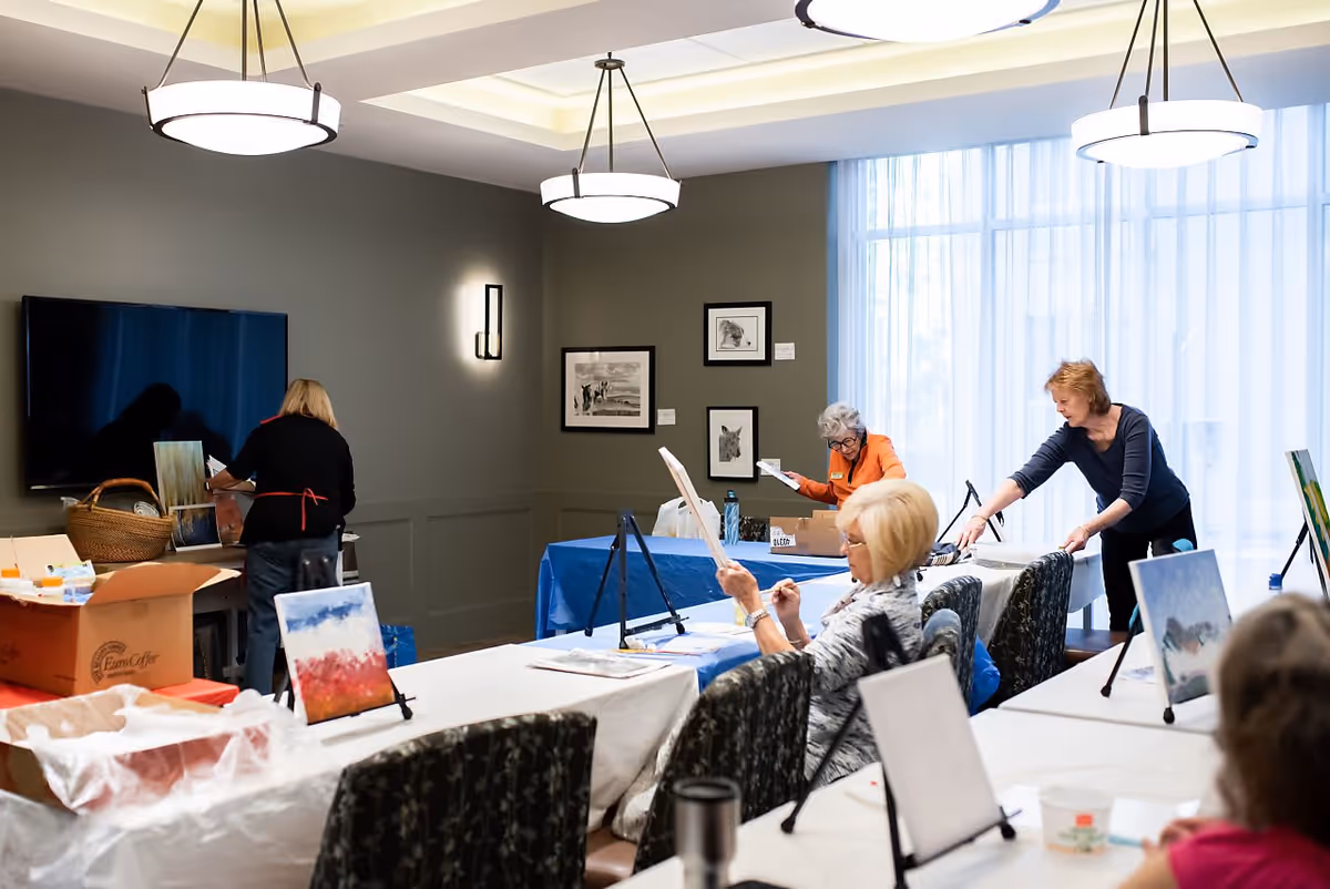 A group of elderly women engaged in a painting activity in a well-lit room with large windows covered by sheer curtains. The room has tables covered with white and blue tablecloths, easels holding canvases, and various art supplies. The walls are decorated with framed artwork, and there are modern ceiling lights providing illumination.
