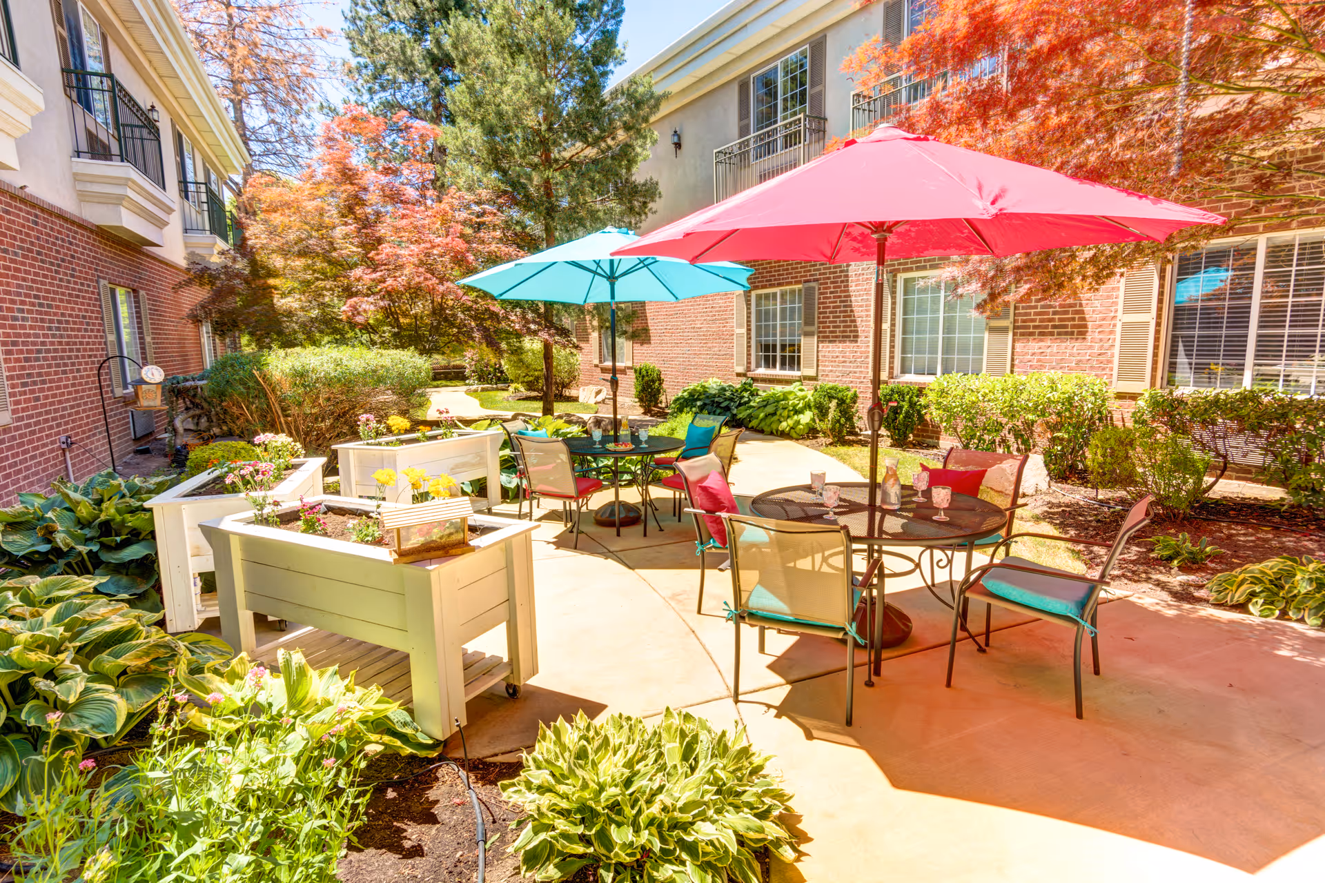 Sunny outdoor courtyard patio with tables, colorful umbrellas, planter boxes, and landscaped gardens beside a brick building.