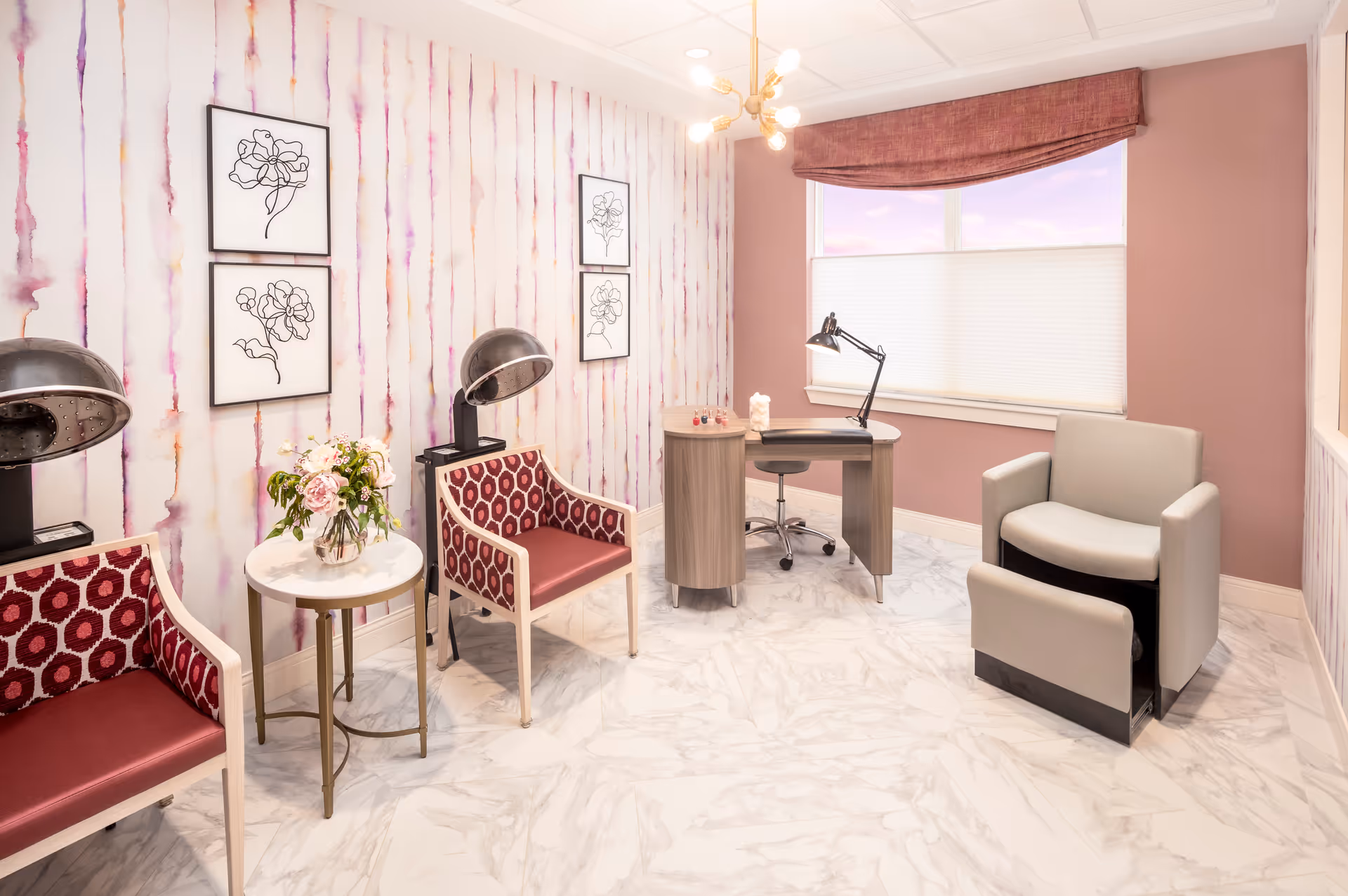 A bright and clean salon room with two vintage hair dryers over red patterned chairs, a small round table with a flower vase, a manicure table with a lamp and chair, and a comfortable beige salon chair. The walls have a pink and white watercolor striped wallpaper with framed floral line art, and a window with a pink valance and white blinds.