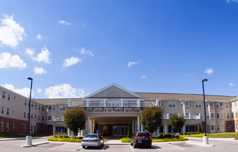 Front exterior view of The Legacy at North Augusta senior living facility with a clear blue sky, two cars parked in front, and well-maintained landscaping including trees and shrubs.
