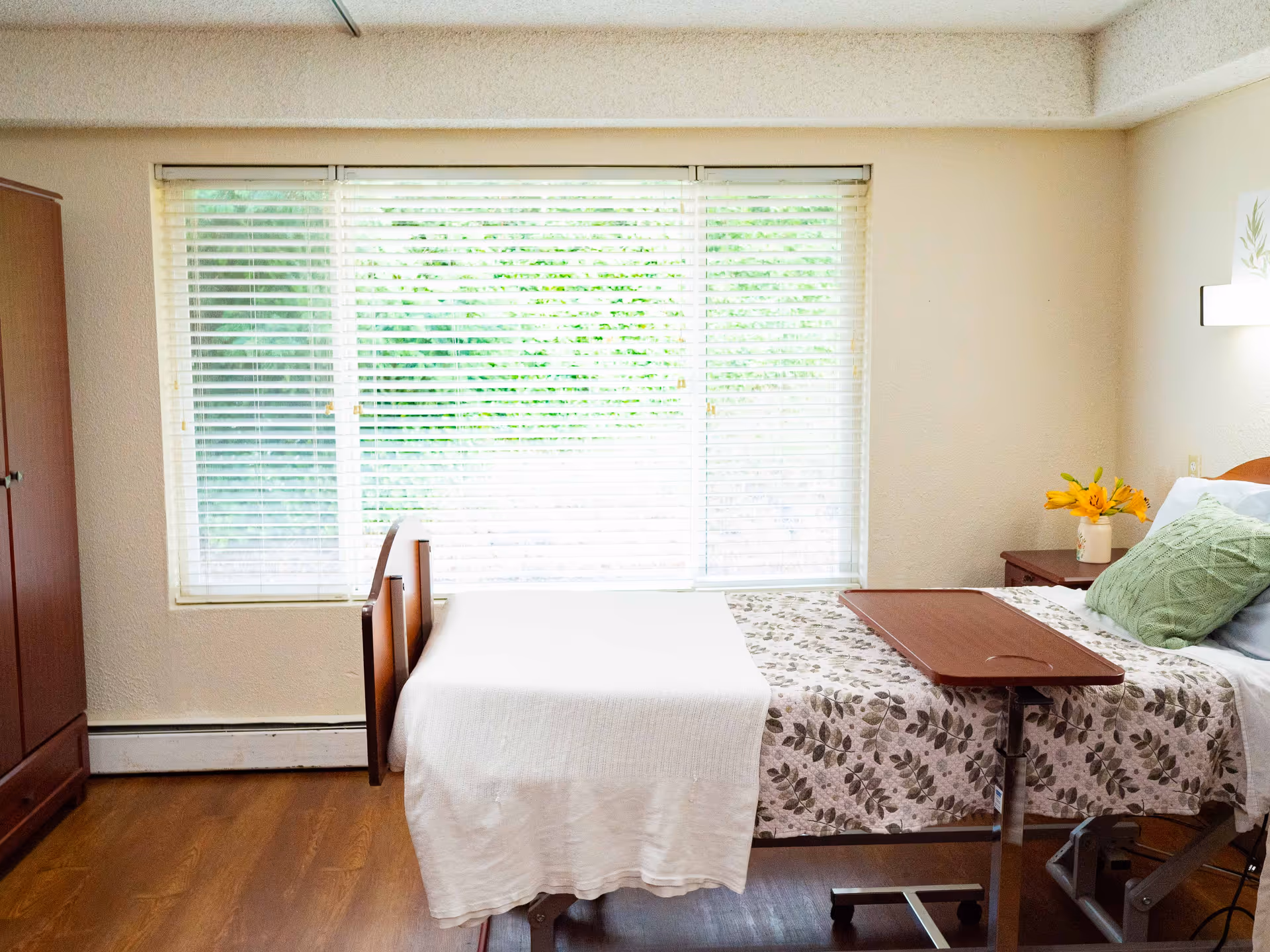 A bright and clean bedroom in a senior living facility featuring a single bed with a floral bedspread and a white blanket. There is a green pillow and a small vase with yellow flowers on the bedside table. A wooden wardrobe stands to the left, and large windows with white blinds allow natural light to fill the room.