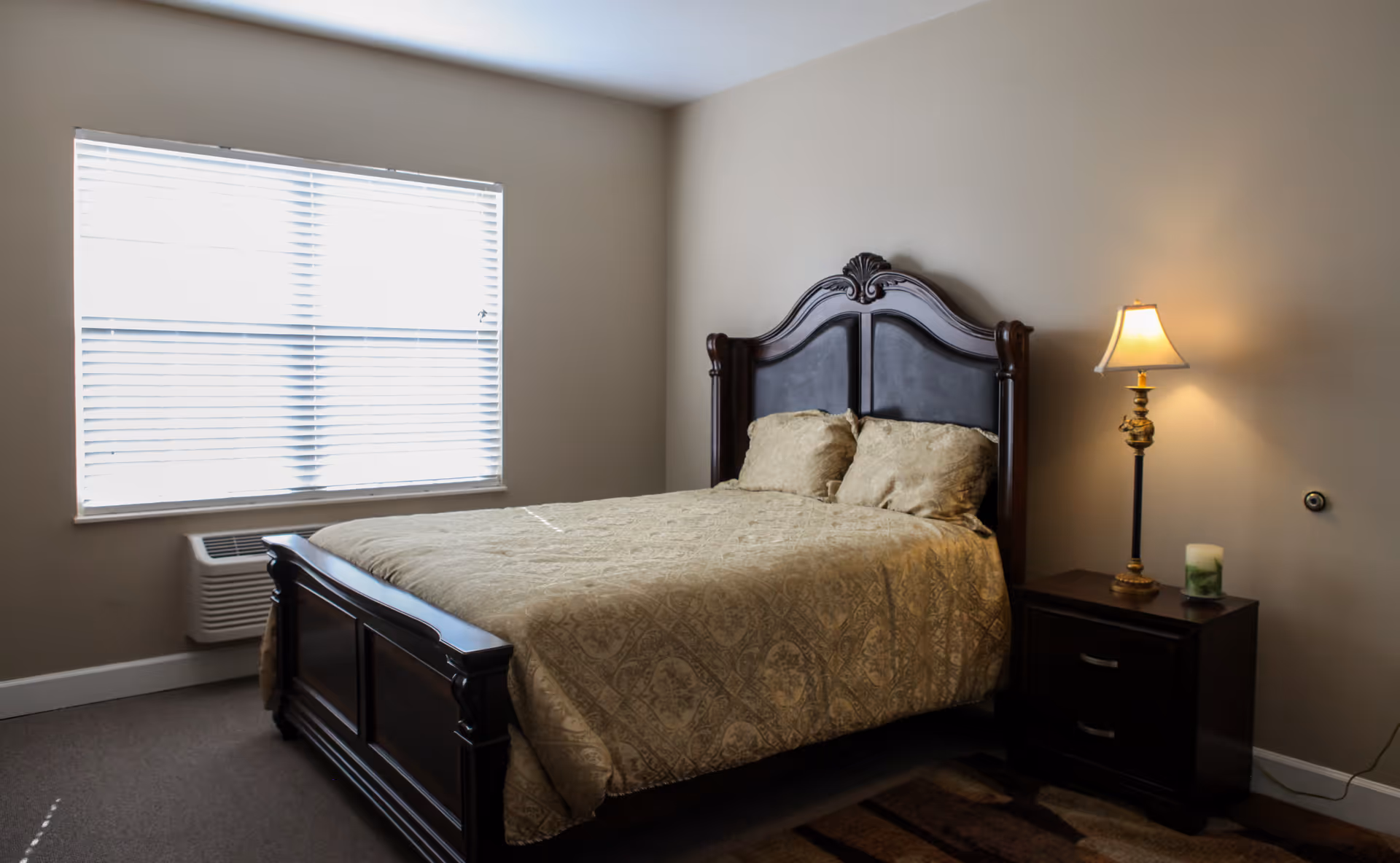 A bedroom with a large wooden bed featuring an ornate headboard, covered with a beige patterned bedspread and two matching pillows. Next to the bed is a dark wooden nightstand with two drawers, a lit table lamp, and a candle. A window with white blinds is on the left wall, and the room has beige walls and carpeted floor.