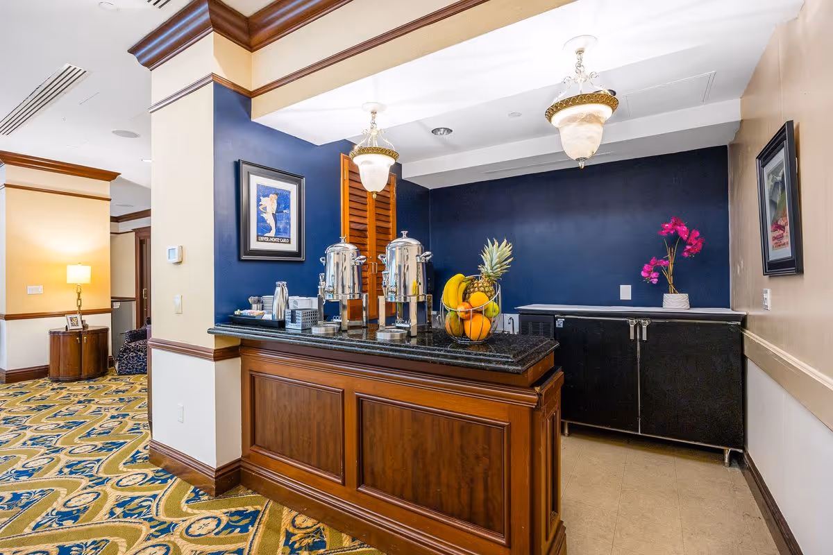 Interior view of a refreshment area in a senior living facility with a wooden counter holding two large beverage dispensers and a bowl of assorted fruits. The walls are painted blue and beige, decorated with framed artwork and a vase with pink flowers. The floor has a patterned carpet and there are ceiling light fixtures above.