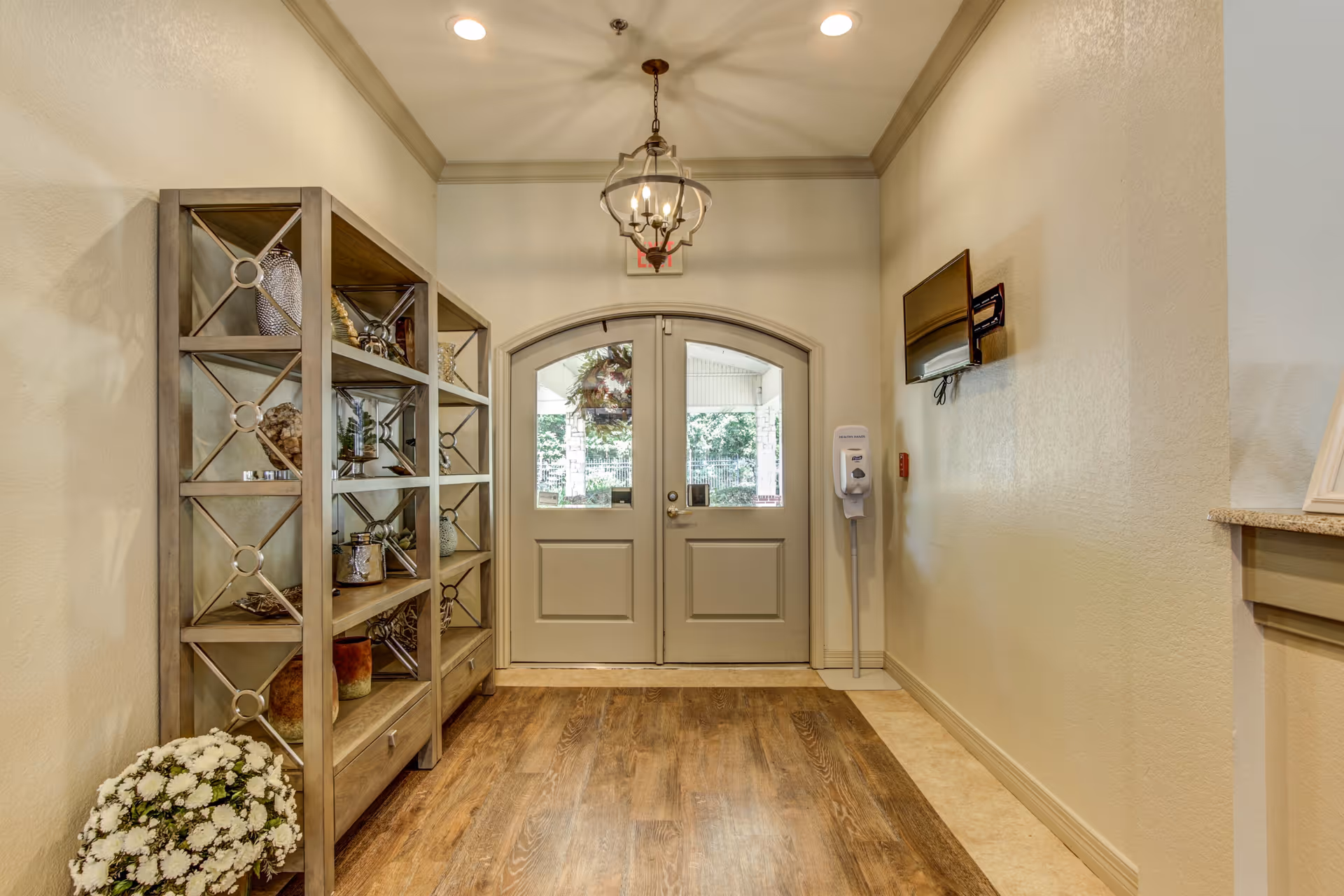 Entry hallway with double doors, a decorative shelving unit on the left, a hanging light fixture, and a wall-mounted hand sanitizer dispenser.