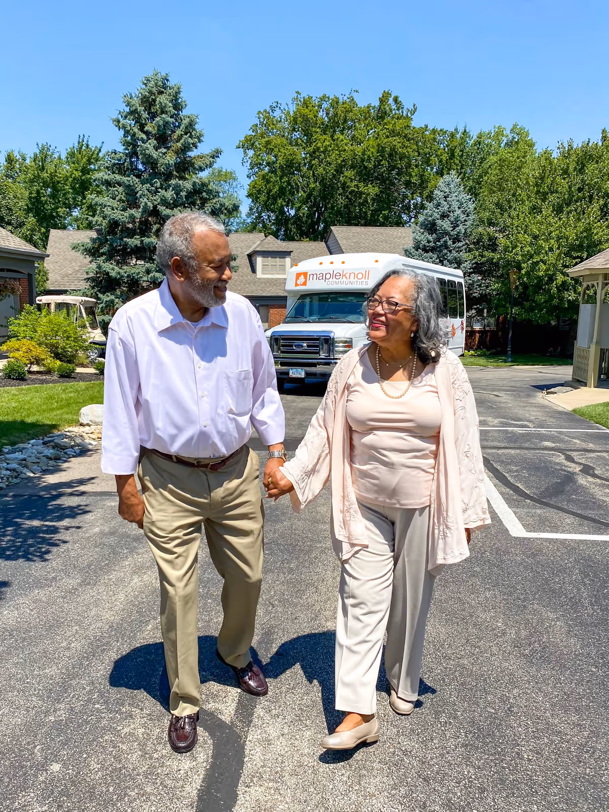 An elderly couple holding hands and walking outside on a sunny day in a residential community. Behind them is a white shuttle bus with the logo and text 'mapleknoll COMMUNITIES'. The area is surrounded by trees and houses.