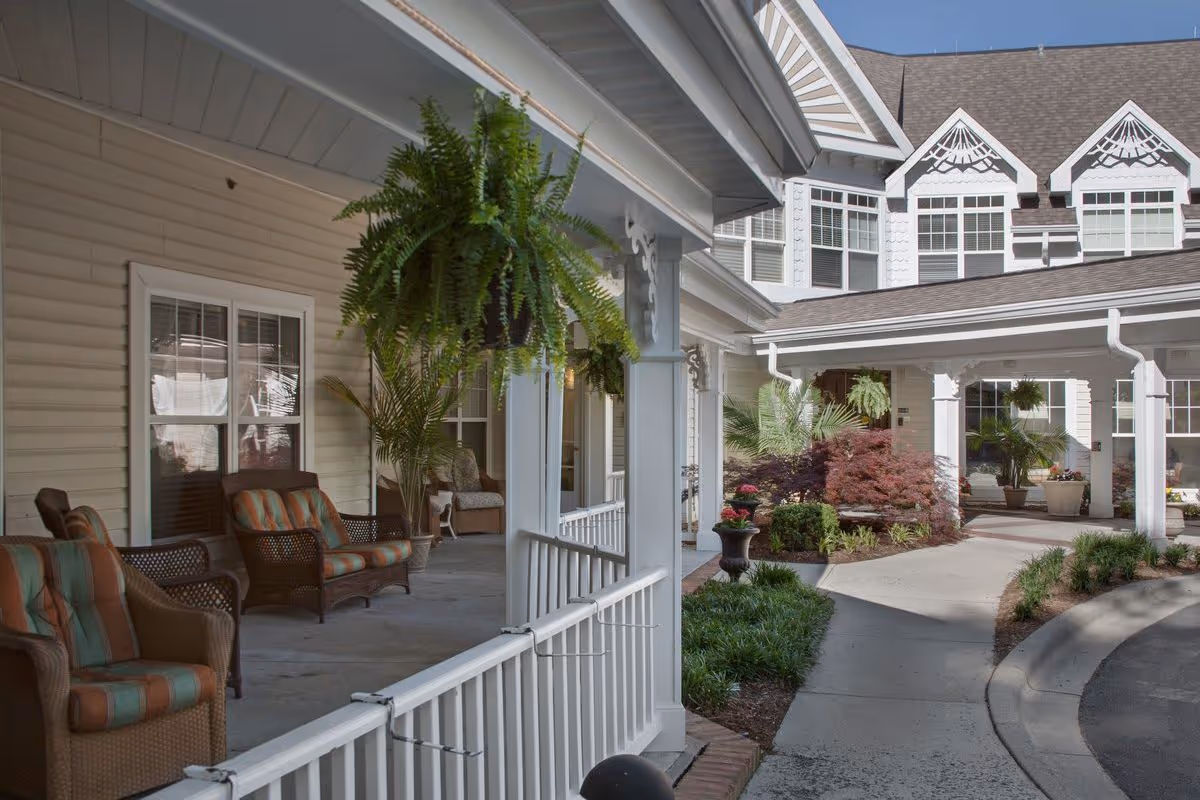 Outdoor covered porch area with cushioned wicker chairs and hanging plants, adjacent to a landscaped garden and the exterior of a multi-story building with white siding and decorative trim under a clear blue sky.