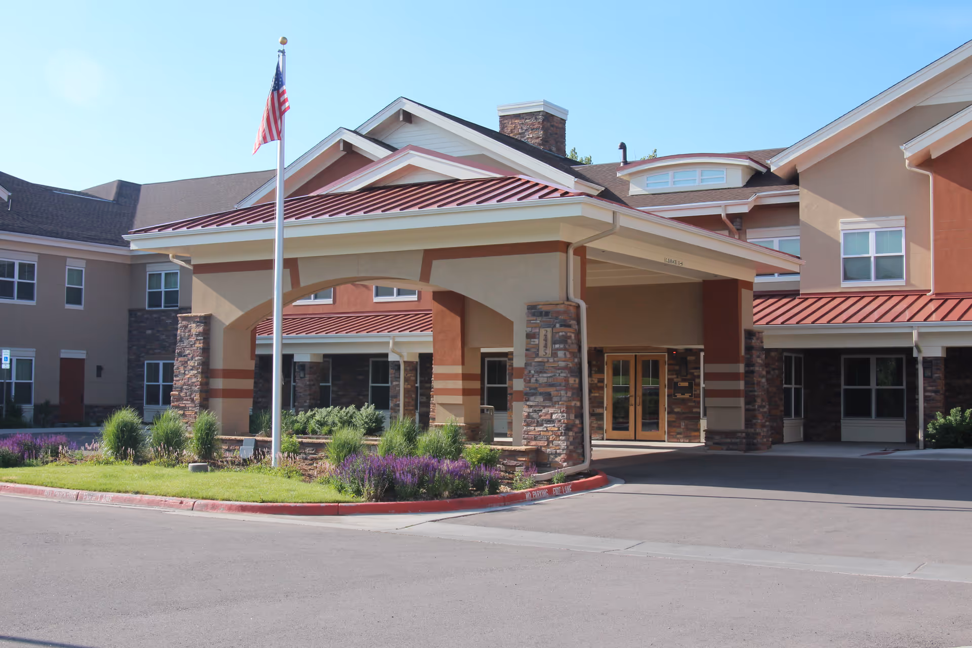 Front entrance of a senior living facility with a covered porte-cochere, American flag, and landscaped driveway.