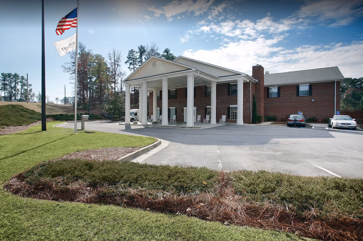 Exterior view of a two-story brick building with white columns at the entrance, several rocking chairs on the porch, two flagpoles with flags, a parking area with a few cars, and landscaped greenery in the foreground under a partly cloudy sky.