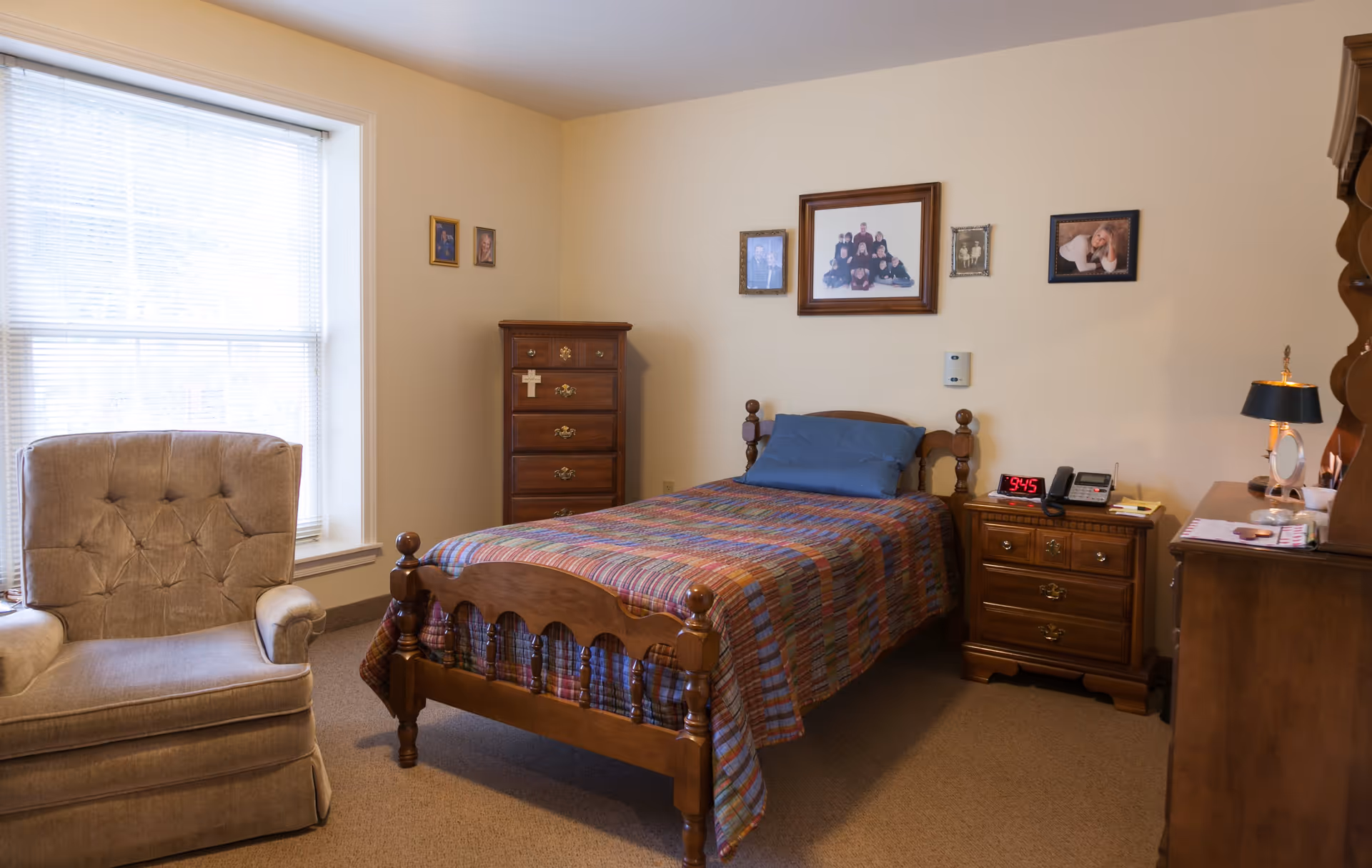 A cozy bedroom with a single wooden bed covered with a colorful plaid quilt and a blue pillow. Next to the bed is a wooden nightstand with a digital clock showing 9:45, a telephone, and a small lamp. There is a wooden dresser with a cross hanging on it and several framed family photos on the beige walls. A cushioned armchair is positioned near a large window with blinds, allowing natural light to fill the room.