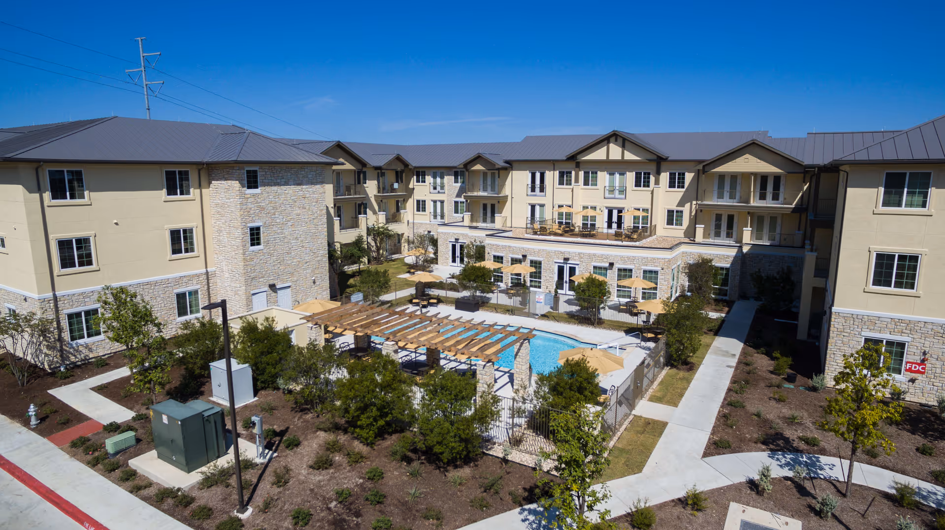 Aerial view of Lake Travis Independent Living facility showing a large, three-story beige building with stone accents surrounding a central courtyard. The courtyard features a swimming pool with lounge chairs and umbrellas, shaded by a wooden pergola. There are landscaped gardens and paved walkways around the building under a clear blue sky.