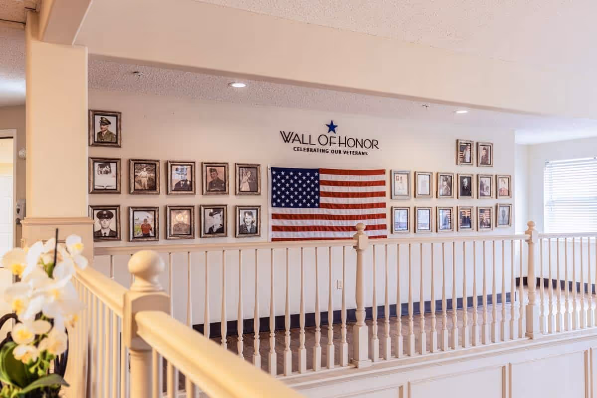 Interior view of a senior living facility hallway featuring a Wall of Honor with framed photos of veterans and an American flag displayed on the wall. The area is well-lit with recessed lighting and has a white railing along the walkway. A potted orchid plant is visible in the foreground.